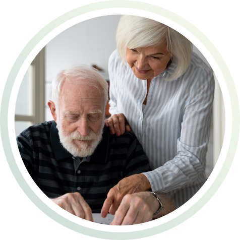 Ein sitzender alter Mann am Tisch, dahinter steht eine Frau und erklärt etwas.