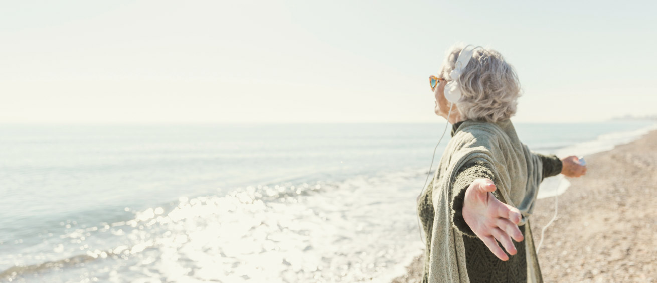 Eine ältere Frau am Strand schaut auf das Meer und streckt die Arme aus.